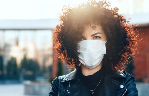Protected caucasian woman with curly hair is posing in a sunny day while looking at camera and wearing a special white mask